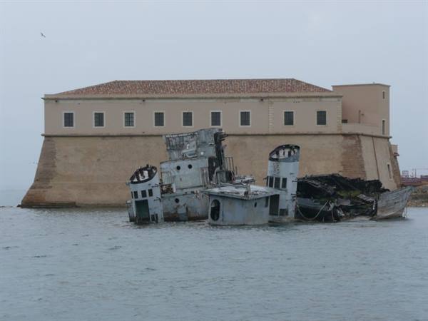 Rusted shipwreck in front of a stone fortress under gray sky