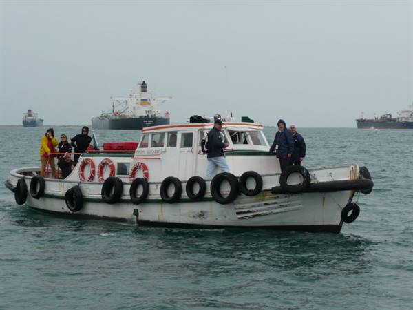 Old white motorboat with tire fenders carries people near oil tankers
