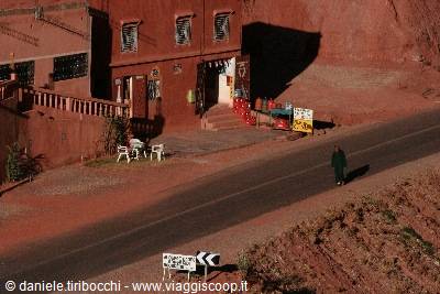 Woman in black walks along the road beside red earthen buildings, bright light