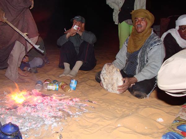 Man in yellow turban sits on sand by the fire, kneading bread dough at night