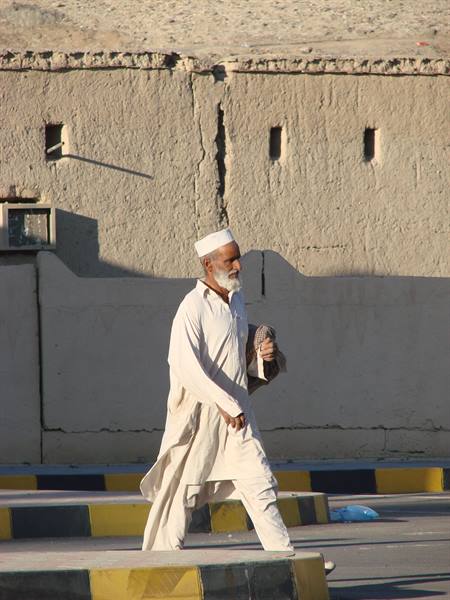 Elderly man in white shalwar kameez and turban walks past an earthen wall
