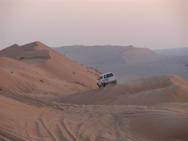 White SUV descends between sand dunes under a pink sky