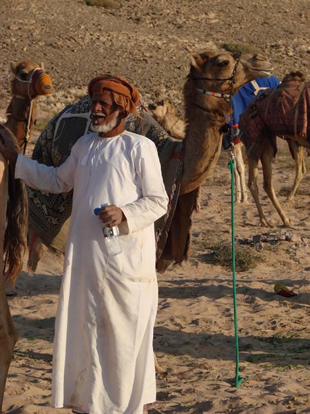 Man in white dishdasha and turban beside saddled camels in the desert