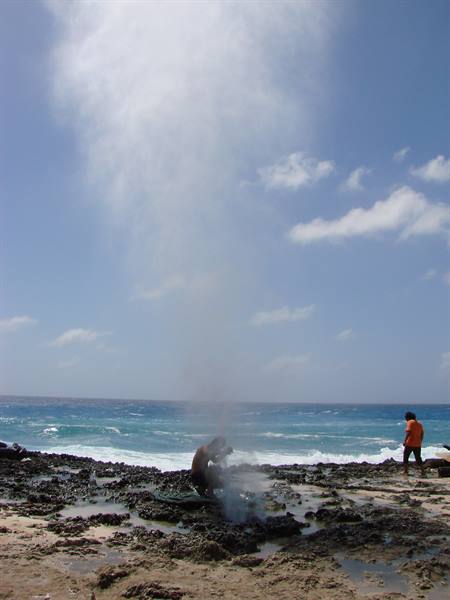Tourist crouches near a blowhole erupting water spray on the black rocky shore