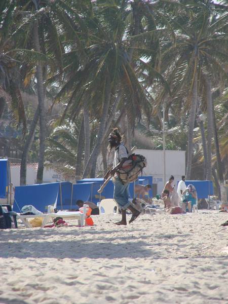 Barefoot man with backpack and long dreadlocks walks past beach chairs and palms