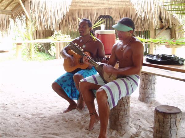Two musicians on wooden stumps play guitar and banjo under thatched hut