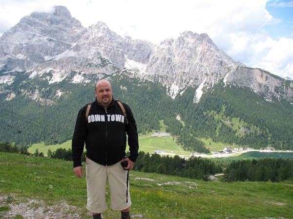 Smiling tourist in black sweatshirt poses on green meadow, Dolomites peaks behind