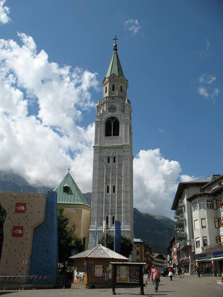 White stone bell tower with green spire in Cortina d'Ampezzo, pedestrians under cloudy sky