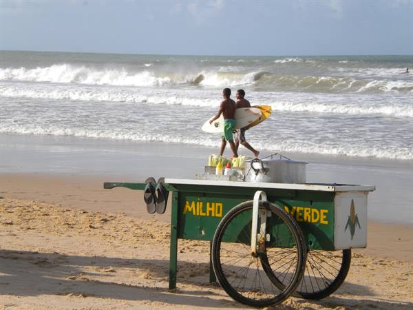 Green corn cart on the sand, two young surfers carrying boards toward the waves