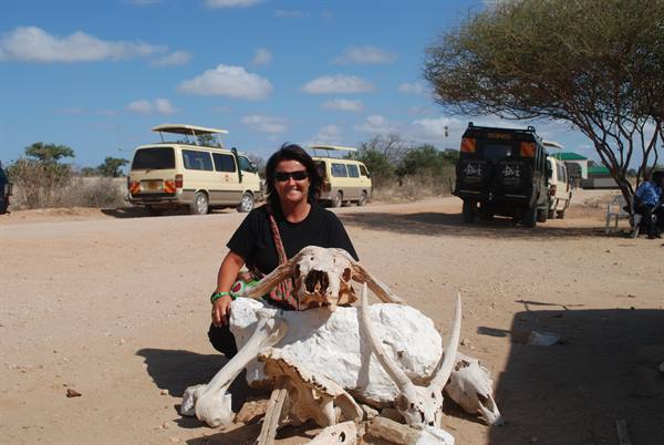 Woman crouching beside animal skulls on sand, safari vans in the background