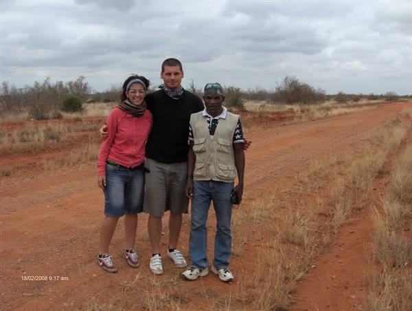 Three friends pose on a red dirt road among dry bushes, cloudy sky