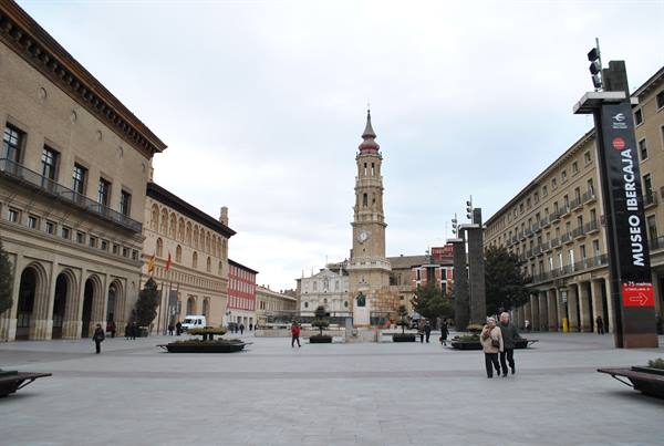 Tourists walk across Plaza del Pilar in Zaragoza, La Seo tower under grey sky