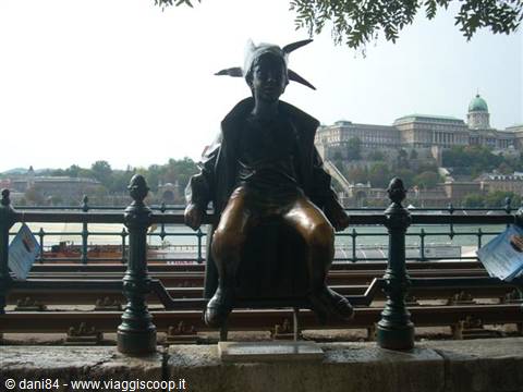 Bronze statue of a child in pointed hat sitting on railing, Buda Castle behind