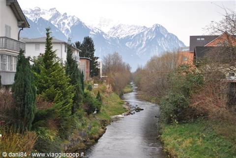 River flowing between houses, snowy Alps behind, evergreens and brick homes