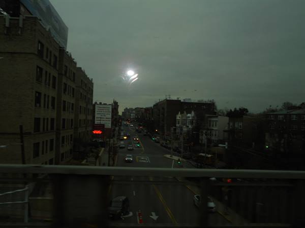 City street with traffic and brick buildings, grey sky, from train window