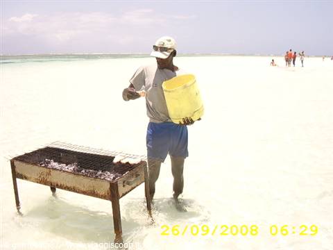 Man with cap and yellow bucket beside a barbecue grill standing in shallow beach water