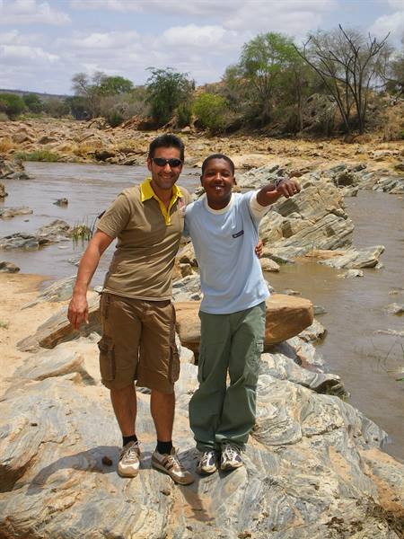 Two smiling men stand on grey rocks beside a muddy river, one pointing ahead