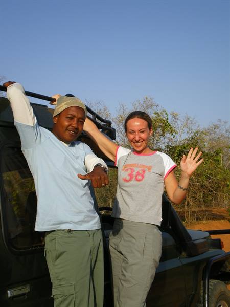 Two people smile beside a black 4x4, surrounded by trees and blue sky