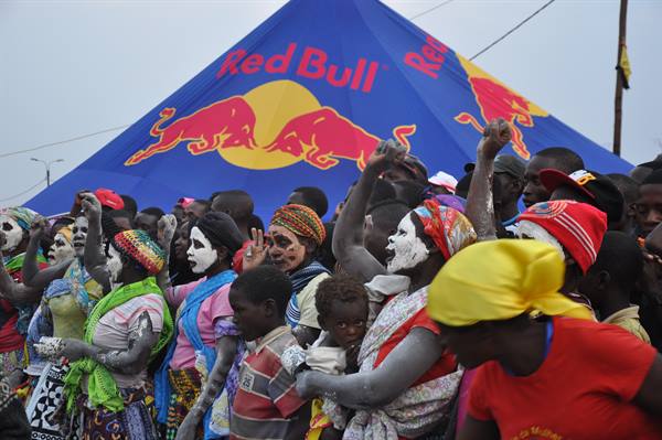 Women with white-painted faces dance in front of a giant Red Bull tent