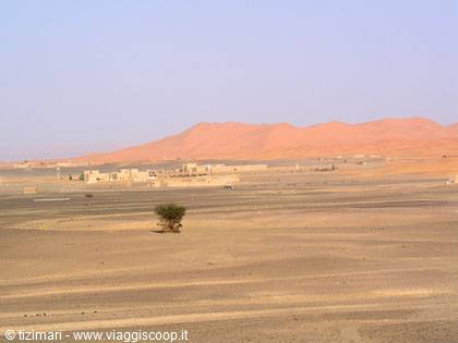 Low village houses at the foot of red desert dunes, lone tree in foreground