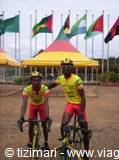 two cyclists in yellow jerseys ride past colorful flags and tents