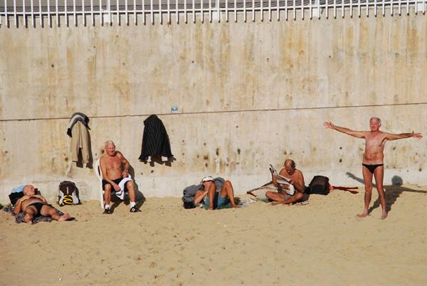 Elderly men sunbathe on sand against concrete wall, one stretching arms wide