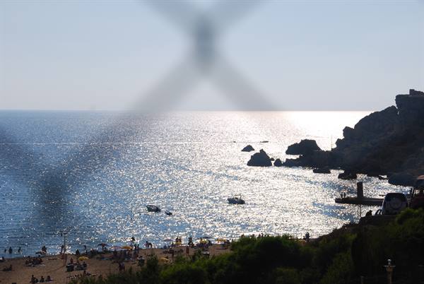 Beach with swimmers and rocks, boats on sea, light through a fence