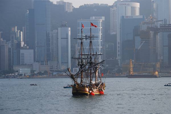 Three-masted tall ship with red flags sails in the bay before Hong Kong skyscrapers