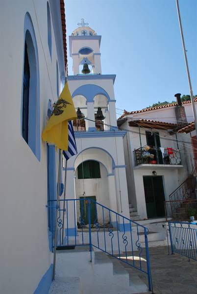 White and blue bell tower of Greek church with yellow flag, houses nearby