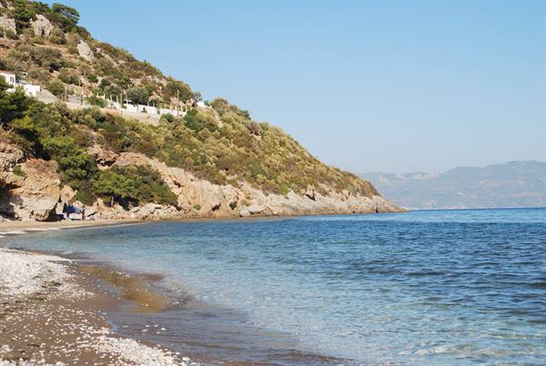 Pebbly beach with turquoise water, green hillside and white houses under clear sky