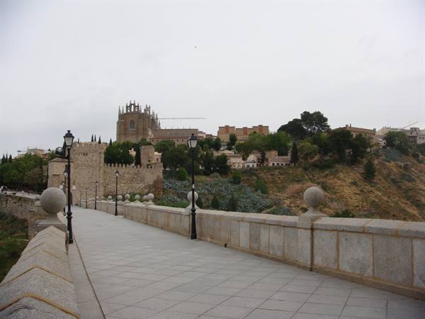 Empty stone bridge with lampposts, ancient walled town on hillside, grey sky