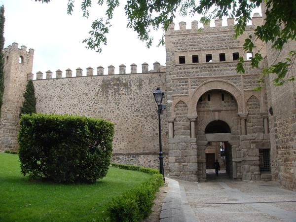 Medieval stone gate with battlements, green lawn, passerby, soft daylight
