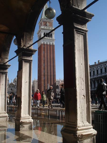 St Mark's bell tower framed by columns, tourists walk on floodwater, bright sunlight