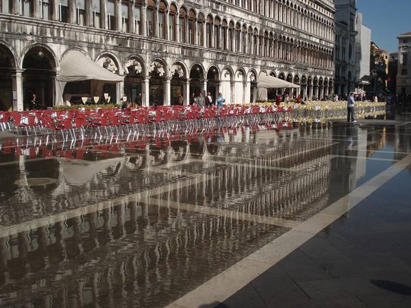 Flooded square with red chairs reflected in water before historic arcades, tourists strolling