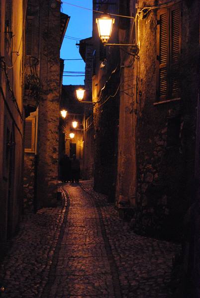 Cobblestone alley at dusk, glowing lamps between old stone houses