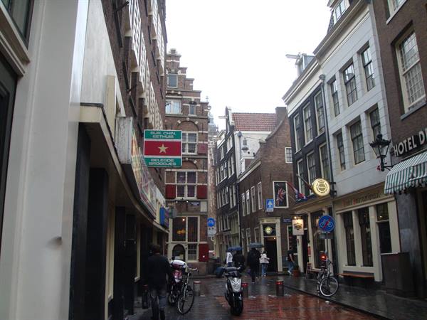 Brick buildings along an Amsterdam alley, cyclists and scooters under grey sky