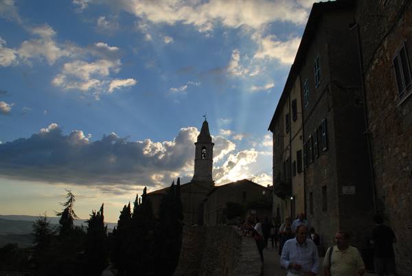 Stone bell tower among old buildings, tourists walk under a cloudy sky