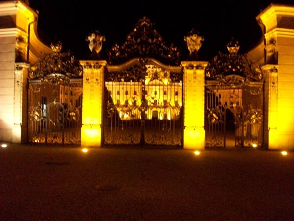 Ornate wrought iron gate lit golden at night before a palace