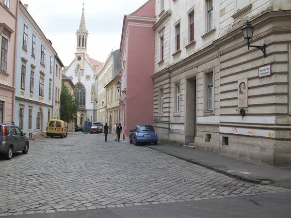 Cobblestone street with parked cars and neo-Gothic church tower between pastel buildings, Bratislava