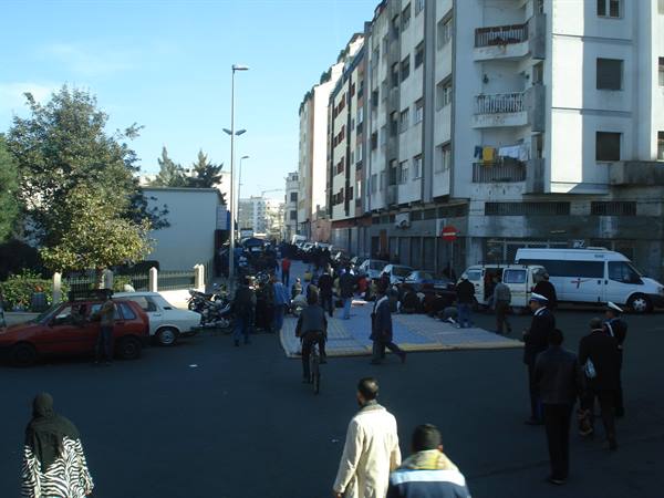 Pedestrians and cars crossing a street lined with white buildings and a van