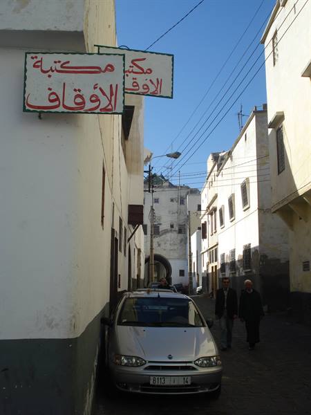 Arabic signs on white wall, silver car and pedestrians in a medina alley