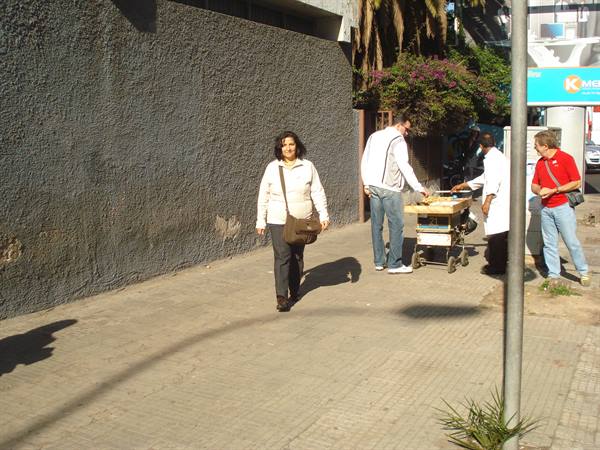 Woman with shoulder bag walks on sidewalk past food cart vendor and people