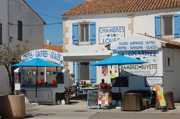 Crepe stand with blue umbrellas in front of white shuttered house, man seated outside