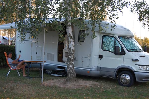 White motorhome parked under a birch tree, man relaxing on orange chair at campsite