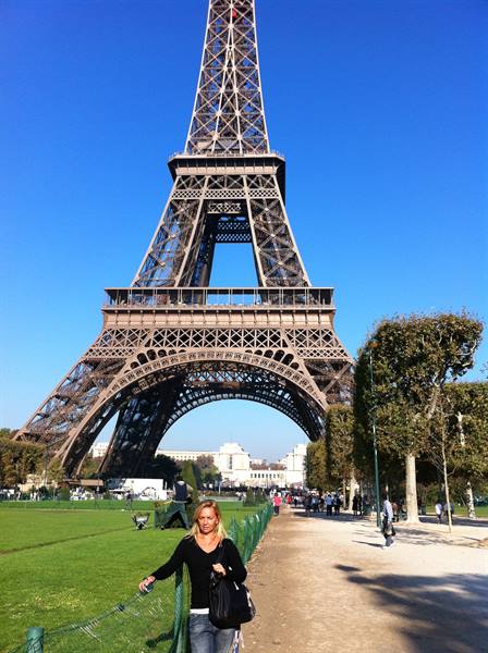 Blonde woman with black bag beside the iron Eiffel Tower, clear blue sky
