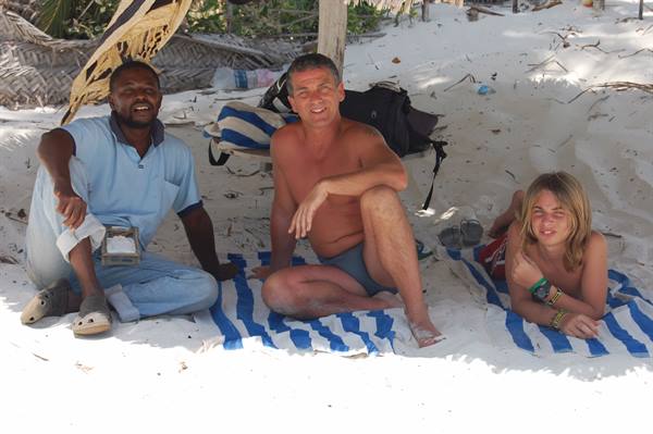 Three people sitting on blue striped cloths under a sandy beach shelter