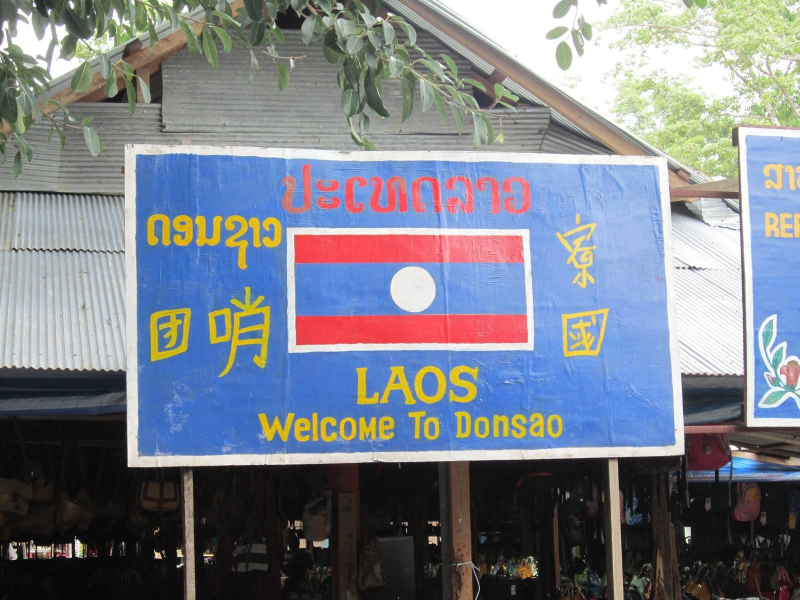 Insegna blu con bandiera del Laos davanti a un mercato di legno a Donsao