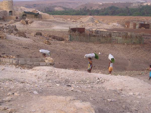Women carrying sacks on their heads walk past stone houses and arid hills