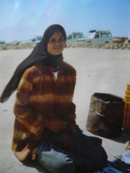 Woman in black headscarf sitting on sand, jeeps parked behind