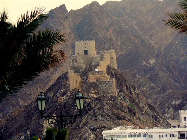 Muttrah Fort atop rocky hill, palm fronds and lampposts in foreground